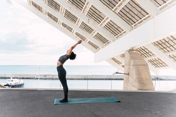 Fit woman performing yoga upward salute pose under a futuristic white metal structure by the ocean © BUDDHA