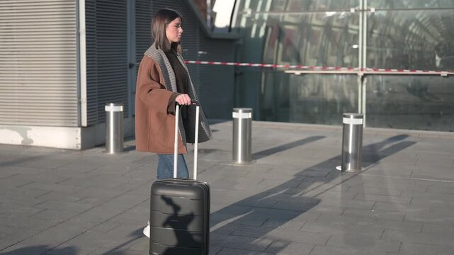 Stylish young woman walking confidently with her rolling luggage outside a modern airport or train station terminal, embarking on a new journey with purpose and independence. Slow motion