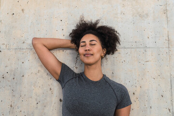 Athletic woman with curly hair and closed eyes resting against a concrete wall, enjoying a moment of peace and mindfulness