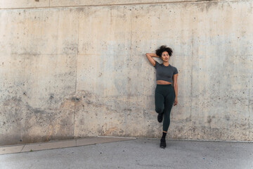 Sporty woman with curly hair and sportswear leaning against concrete wall after outdoor workout