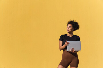 Young woman is holding a laptop and looking away while standing against a yellow wall