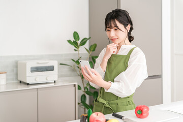 A young Asian woman wearing an apron cooking in the kitchen while looking at her smartphone