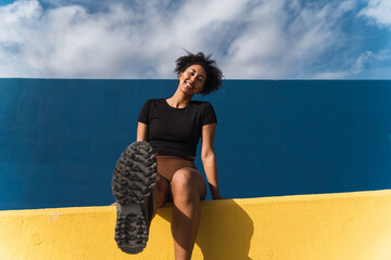 Smiling young woman showing sole of shoe while sitting on colorful wall under blue sky