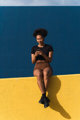 Young woman wearing sportswear using her smartphone sitting on a low wall with a blue and yellow background