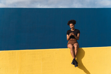 Smiling young woman using mobile phone while sitting on colorful wall with blue sky on background