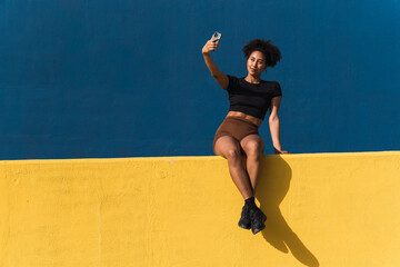 Smiling young woman taking a selfie with her smartphone while sitting on a yellow wall with a blue background