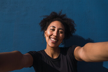 Young smiling woman taking a selfie with outstretched arms against a blue wall background