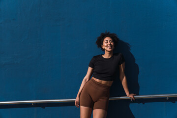 Smiling young woman with brown shorts and black t-shirt leaning on a railing in front of a blue wall