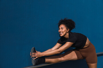 Smiling sportswoman stretching legs against blue wall, enjoying her outdoor workout