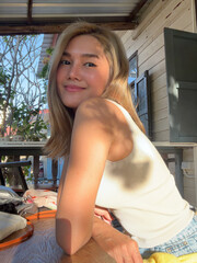 Asian woman with blonde hair smiling and sitting  at table, relaxed and confident posing with warm sunlight.