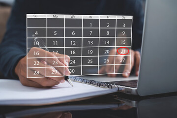 Person planning schedule and marking an important date on a digital calendar overlay while writing notes in a notebook next to a laptop, highlighting time management and event planning.