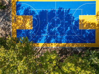 Top-down aerial view of an outdoor basketball court surrounded by trees. Bright blue and yellow surface with strong shadows creates a graphic, geometric composition. Sports, urban recreation, active