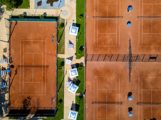 Top-down aerial view of clay tennis courts separated by a walkway with umbrellas and seating. Clean geometric layout, symmetry, warm colors, outdoor sports and recreation concept