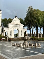 Obraz premium The Telsiai Cathedral complex in Lithuania features a classic white archway adorned with a cross, flanked by sculpted niches and statues. In front, a stone fountain with a central pedestal 