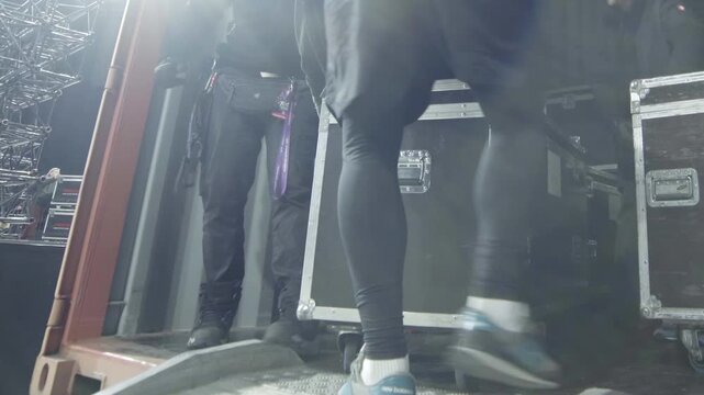 Workers unload truck trailer with various musical instruments and equipment cases on stage closeup. Movers work with band supplies indoors