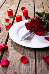 Fresh red roses and cutlery in plate by candles on wooden table, copy space
