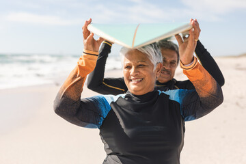 Happy senior multiracial woman carrying surfboard over head with man at sunny beach during weekend