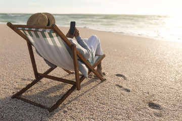 Senior woman sitting on chair using mobile phone while relaxing at beach during sunset
