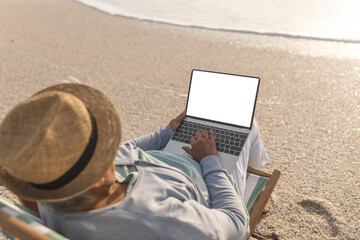 senior woman wearing hat using blank screen laptop with copy space at beach during sunset