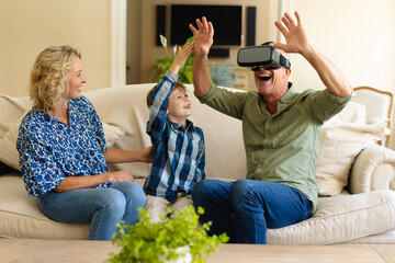 Happy caucasian grandmother and grandson watching grandfather wearing vr headset on couch at home