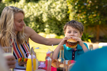 Blond caucasian mother watching son eating burger in the garden with family