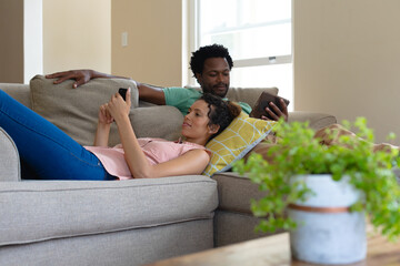Young multiracial couple using smartphone and tablet pc while relaxing on sofa at home
