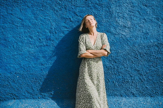 Relaxed woman in green dress leaning against blue wall in sunlight