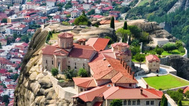 Cinematic aerial orbit of St. Stephen's Monastery in Meteora, Greece. Drone footage reveals the stunning Orthodox complex, gardens, and red roofs against the dramatic cliffs