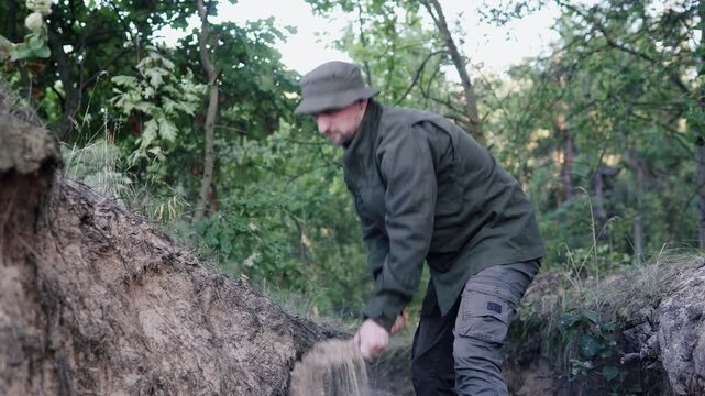 A soldier in camouflage digs a trench with a shovel in a dense woodland, forming a foxhole or dugout for protection from enemy fire.