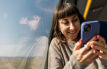 A cheerful young woman texting on a mobile phone on a train. Side view of a woman on a train. A girl using social media.