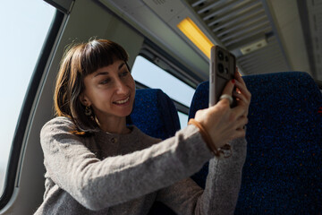 A train passenger enjoys the journey. A happy woman takes photos on the train with her smartphone. She creates content for social media.