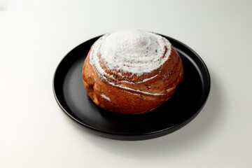 Round, delicious bun with cinnamon and powdered sugar on a black round plate on a white background