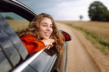 A happy woman leans out the car window, feeling free and enjoying the fresh air at sunset. A traveler on a road trip surrounded by agricultural fields. Concept of travel, freedom, and fun. © maxbelchenko