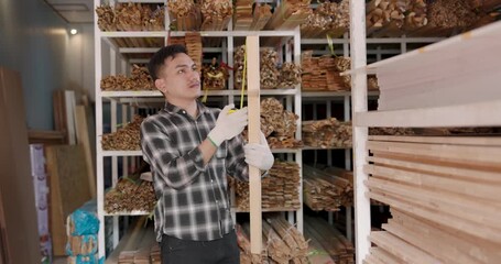 Asian adult male professional tradesman holding long wooden plank upright, checking length balance surface quality while working inside wood supplier business operation warehouse