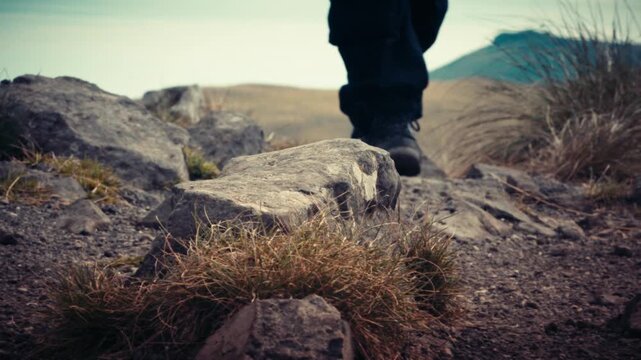 Close up detail of hiking boots traversing rocky ground covered in dry grass. Focus on the texture of stones and the action of walking through rugged outdoor terrain.