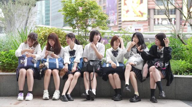 A group of young women are sitting on a bench, posing for a picture. They are wearing school uniforms and have backpacks with them. Scene is casual and friendly