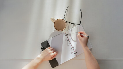 Person's hands writing notes in a planner next to a cup and glasses on a minimalist desk, top view