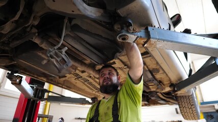 Adult professional repairman fixing car at service or garage. Male auto mechanic works underneath a lifting vehicle at workshop. Concept of regular automobile maintenance. Close up Slow motion