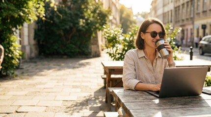 Smiling adult Caucasian freelancer in sunglasses drinking from a thermos while typing on a laptop at an outdoor cafe table with warm golden hour sunlight.