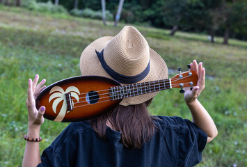 A person in a straw hat holding a palm-designed ukulele outdoors: a serene scene in a grassy field with a person wearing a wide-brimmed straw hat, holding a wooden ukulele adorned with a palm tree pat
