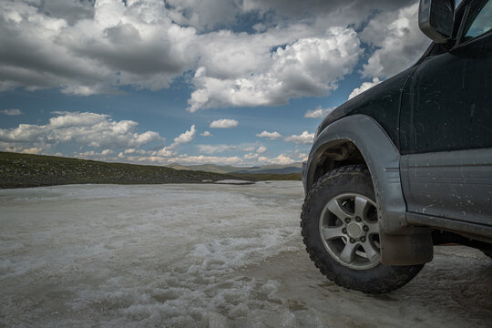 The left front section of an off-road vehicle is parked on an unmelted ice sheet in a permafrost region. A medium shot focuses on the aggressive tire tread against thawing ground.