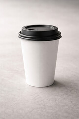 A plain white disposable paper coffee cup with a black plastic lid stands upright on a light grey surface.