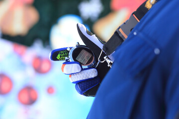 A close-up of a gloved hand holding a smartwatch. The background features colorful holiday decorations, suggesting a festive winter atmosphere.