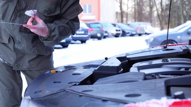A man in a green jacket checks the oil level in a car engine during winter. Snow covers the ground, and parked cars are visible in the background.