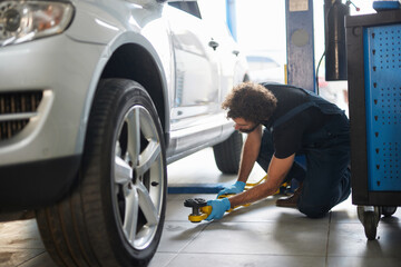 A skilled car mechanic crouches beside a vehicle, using equipment to fix a tire in a well-equipped workshop filled with tools and machinery during daytime.