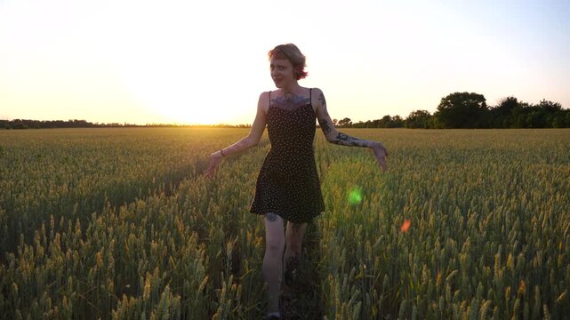 Cheerful punk girl in short black dress fooling around while walks among wheat meadow. Happy female hipster with tattoos having fun during goes through green barley field at sunset. Freedom concept