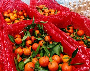 Fresh tangerines with green leaves overflow vibrant red plastic, market freshness. Ripe mandarin oranges in red mesh bags with leaves, golden champagne next to them, bright produce display.