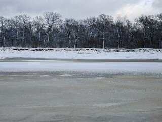 Frozen lake under stormy winter sky. Vast icy lake surface with snow patches, dense bare forest horizon and dramatic dark clouds overhead.