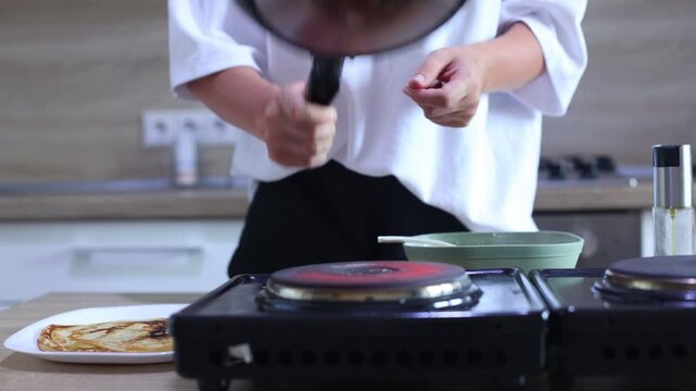 A person expertly flips a golden-brown pancake in a skillet using a flick of the wrist, then slides it onto a plate with other finished pancakes, ready to serve.
