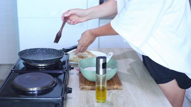 A person prepares to cook another pancake by greasing a hot skillet, pouring in batter, and swirling it evenly across the pans surface.